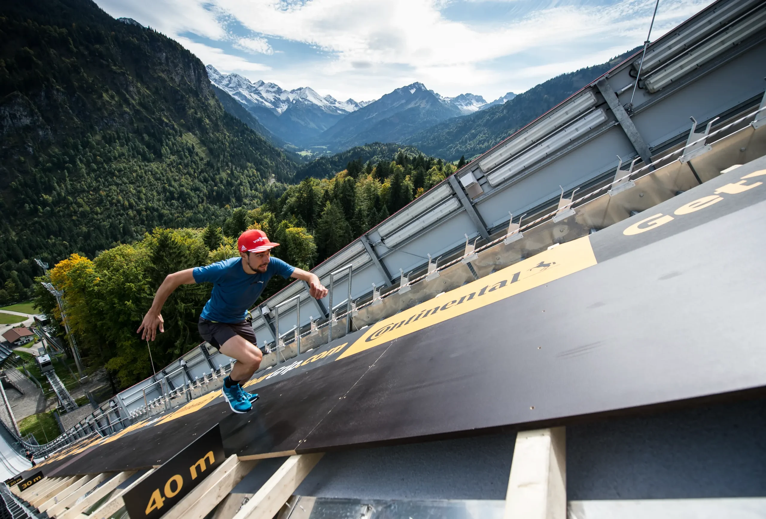 Ein Sportler läuft eine schwarze Fläche auf einer Skisprungschanze im Sommer hoch. Im Hintergrund sind Wald und Berge zu sehen.