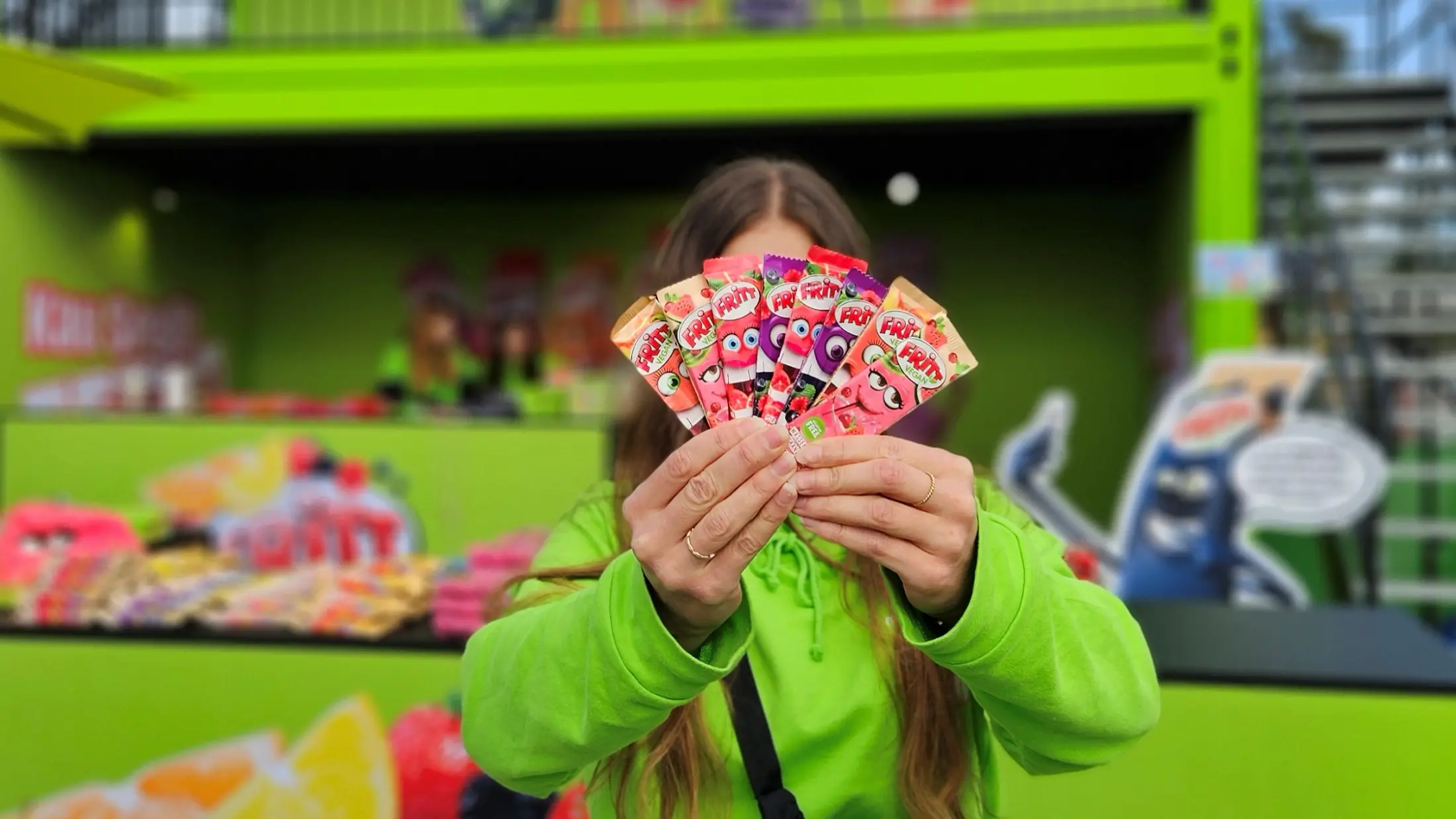 Promoterin hält FRITT-Produkte vor ihr Gesicht. Der FRITT-Stand ist im Hintergrund unscharf zu sehen.