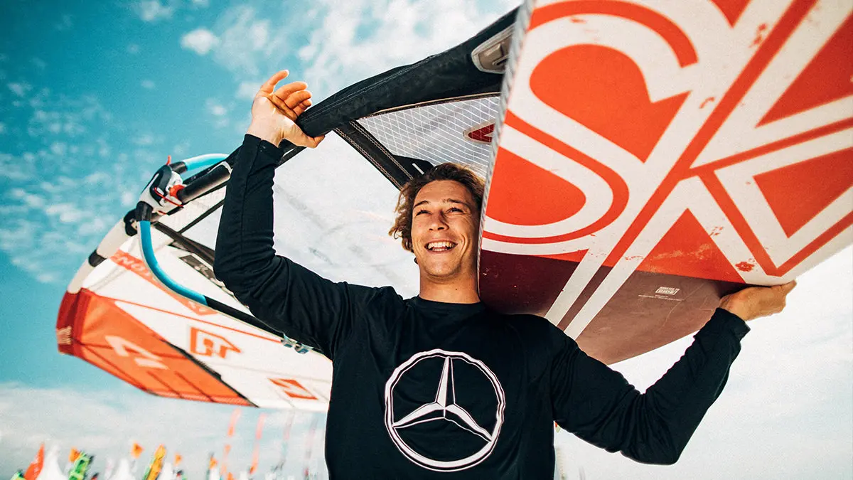 Mann trägt ein Windsurfbrett auf der Schulter. Das Segel ragt über seinen Kopf. Auf seinem Langarm-Shirt ist das Mercedes-Benz-Logo abgebildet.