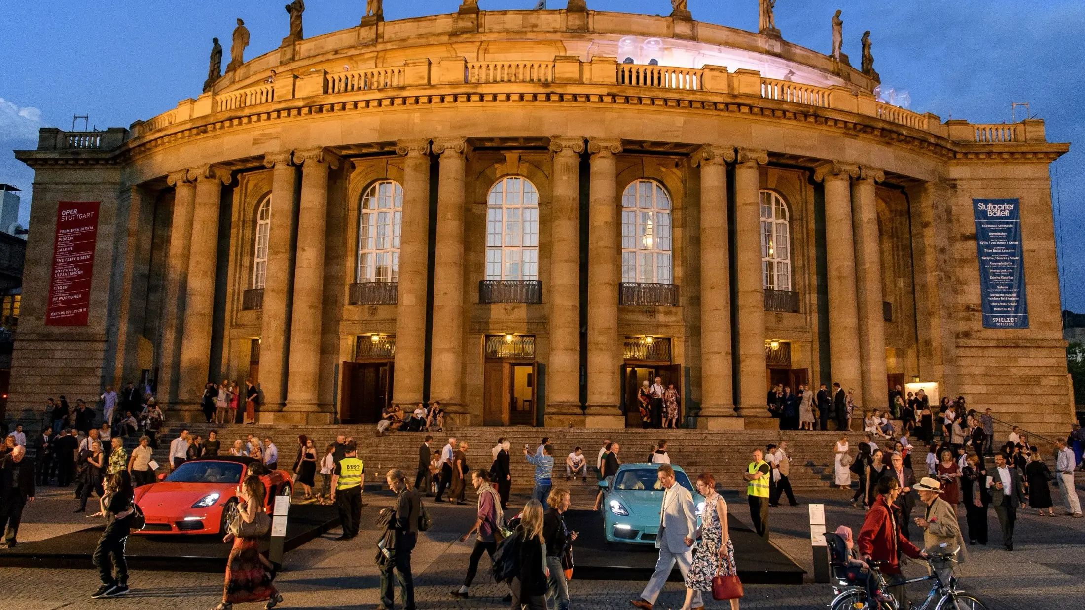Die Stuttgarter Oper von außen in den Abendstunden, davor stehen ein roter und ein blauer Porsche. Menschen gehen vorbei und kommen und gehen in das Gebäude.
