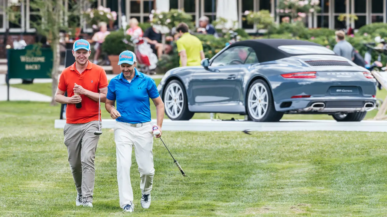 Zwei Männer in bunten Polo-Shirts laufen mit Golfschlägern über den Golfrasen. Im Hintergrund steht ein Porsche und Menschen sitzen auf einer Terrasse.