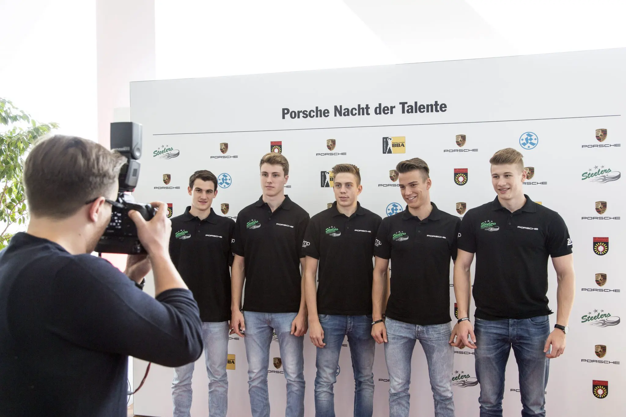 Fünf junge Männer in einheitlichen schwarzen Poloshirts von Porsche stehen vor einer Logo-Wand der "Porsche Nacht der Talente". Die jungen Männer werden von einem Fotografen fotografiert.
