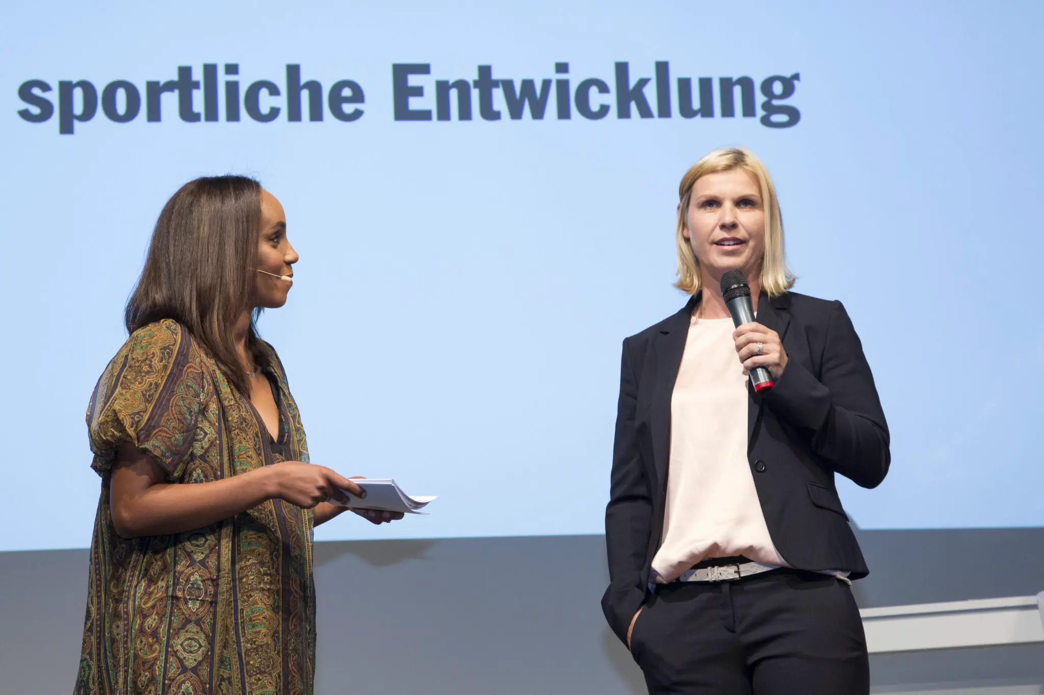 Zwei Frauen stehen auf der Bühne der Porsche "Nacht der Talente" im Hintergrund ist auf einer Leinwand "sportliche Entwicklung" zu lesen.