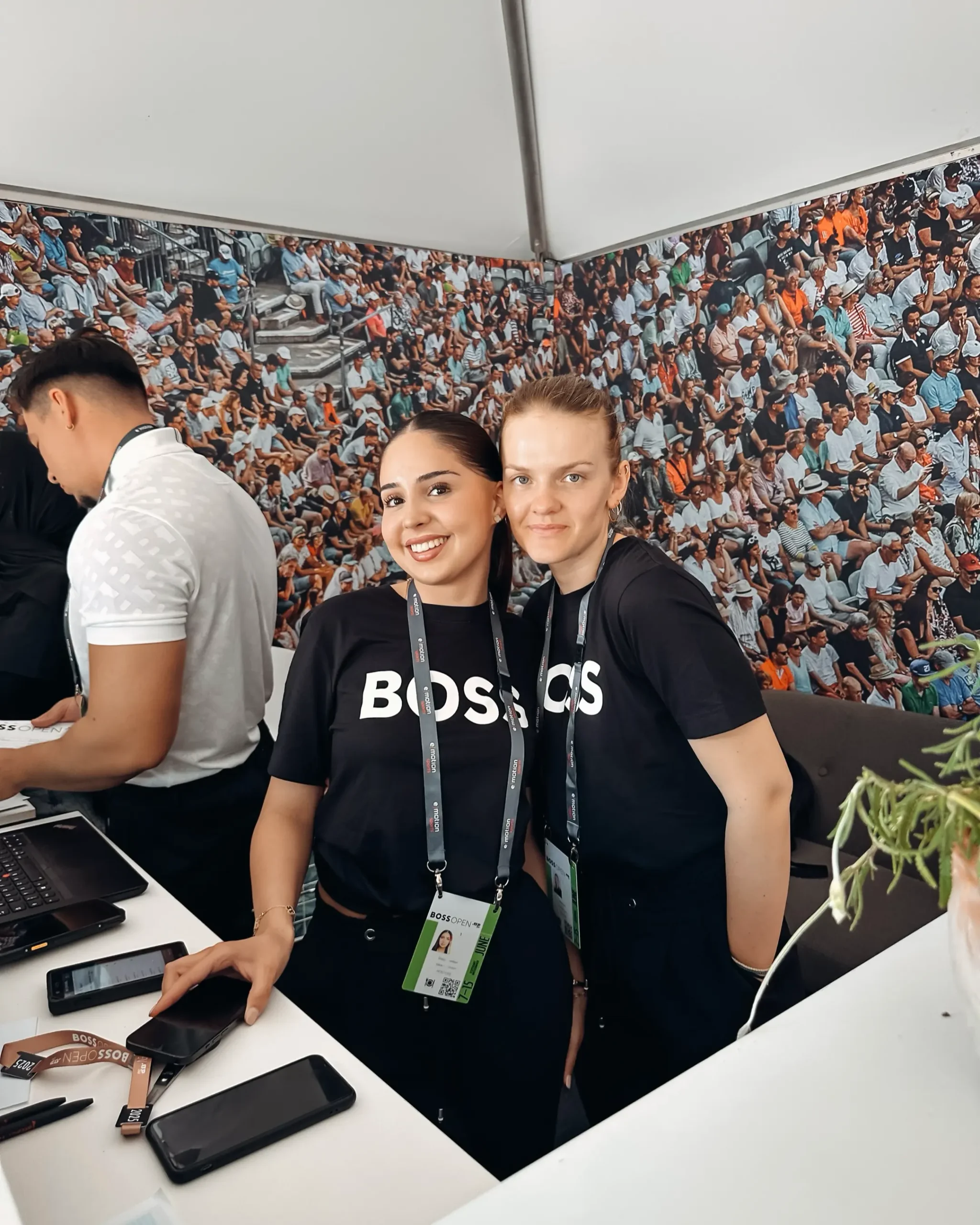Zwei Frauen in schwarzen T-Shirts mit der Aufschrift "BOSS" und ein Mann im weißen T-Shirt stehen hinter einer Registrierungs-Theke. Im Hintergrund ist eine Fototapete, die Zuschauer der BOSS OPEN zeigt.