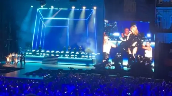 Bühne bei Nacht mit blauen Lichteffekten, auf einer Videoleinwand ist Kylie Minogue mit Tänzern und der Band zu sehen.