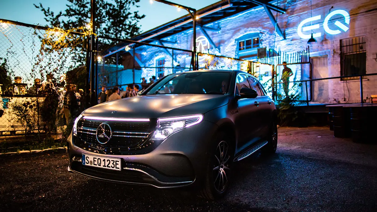 Ein Mercedes-Benz EQ Auto steht vor einer eingezäunten Event-Location der EQ Night. Das Gebäude ist neonfarben angestrahlt und hinter dem Zaun stehen Menschen.